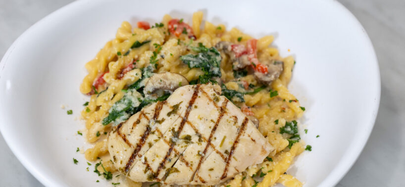 Grilled chicken breast served on a bed of pasta with vegetables and herbs, topped with grated cheese on a white plate.