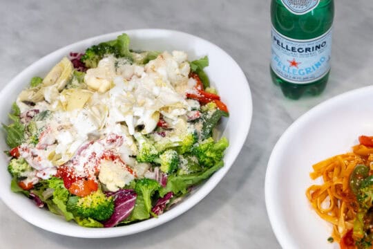 A bowl of mixed salad with assorted vegetables and cheese, a plate of pasta with tomato sauce, and a bottle of San Pellegrino on a marble surface.