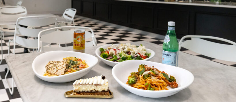 Three plates of pasta and salad, a slice of cake, a can of orange soda, and a bottle of sparkling water on a marble table in a modern, empty restaurant.
