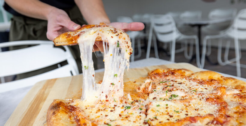 A person lifts a cheesy slice of pizza from a wooden board, stretching melted cheese, with empty white chairs and tables in the background.