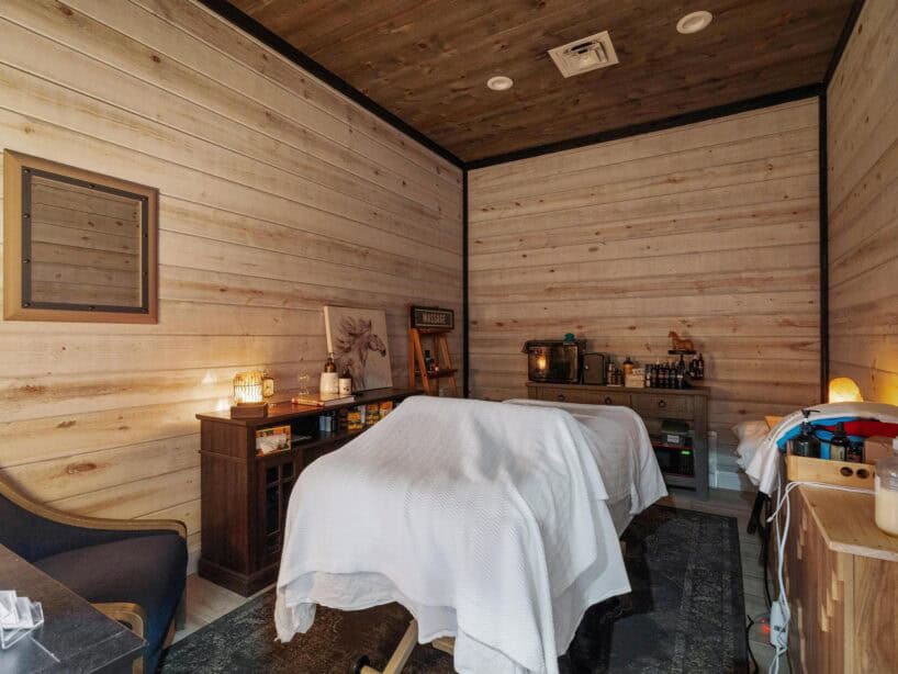 A spa treatment room with two massage tables covered in white sheets, wooden walls, dim lighting, and various spa supplies on shelves and counters.