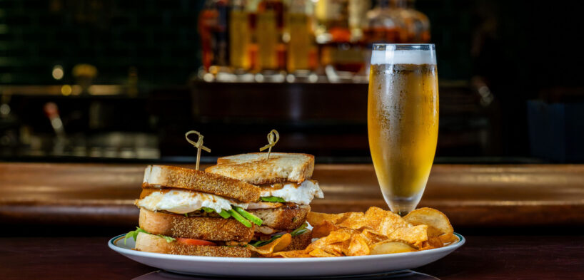 A plate with two stacked sandwiches and potato chips is served next to a tall glass of beer, with bottles blurred in the background.