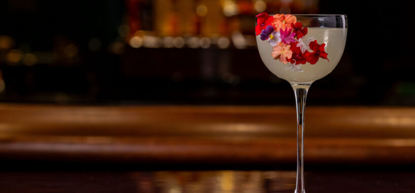 A cocktail in a coupe glass is garnished with colorful edible flowers, with additional flowers scattered on the bar surface in front of it.
