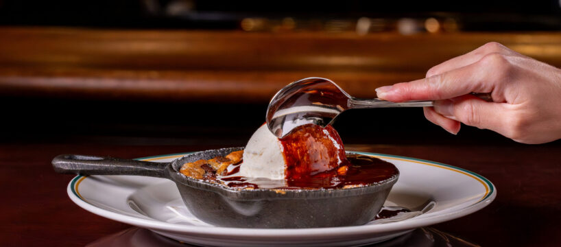 A hand scoops vanilla ice cream topped with chocolate sauce from a skillet dessert on a white plate.