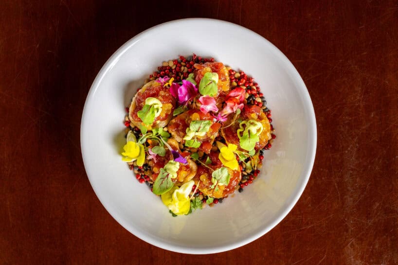 A white bowl filled with a colorful dish of plated vegetables, edible flowers, herbs, and grains, set on a dark wooden surface.