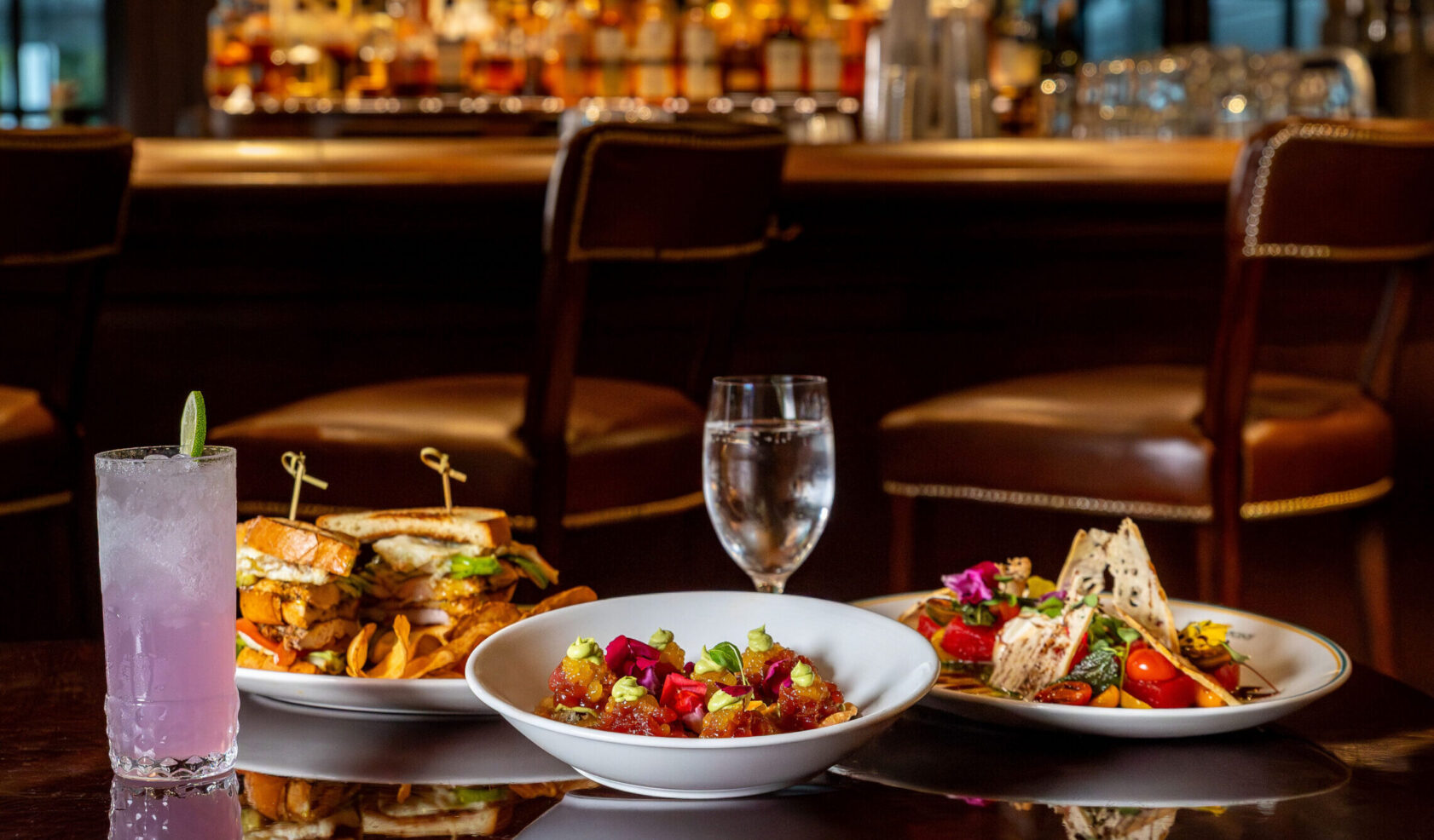 Three plates of assorted food and a pink drink are arranged on a table in front of a bar with shelves of bottles and empty bar stools in the background.