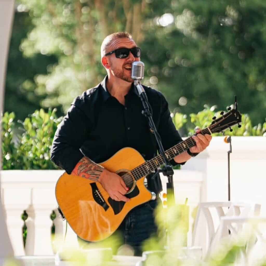 A man wearing sunglasses and a black shirt plays an acoustic guitar and sings into a microphone outdoors.
