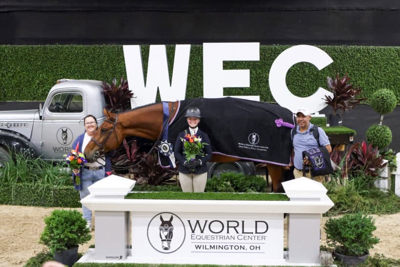 Three people and a horse pose with flowers and a prize ribbon in front of a World Equestrian Center sign in Wilmington, Ohio. A vintage truck and large WEC letters are in the background.