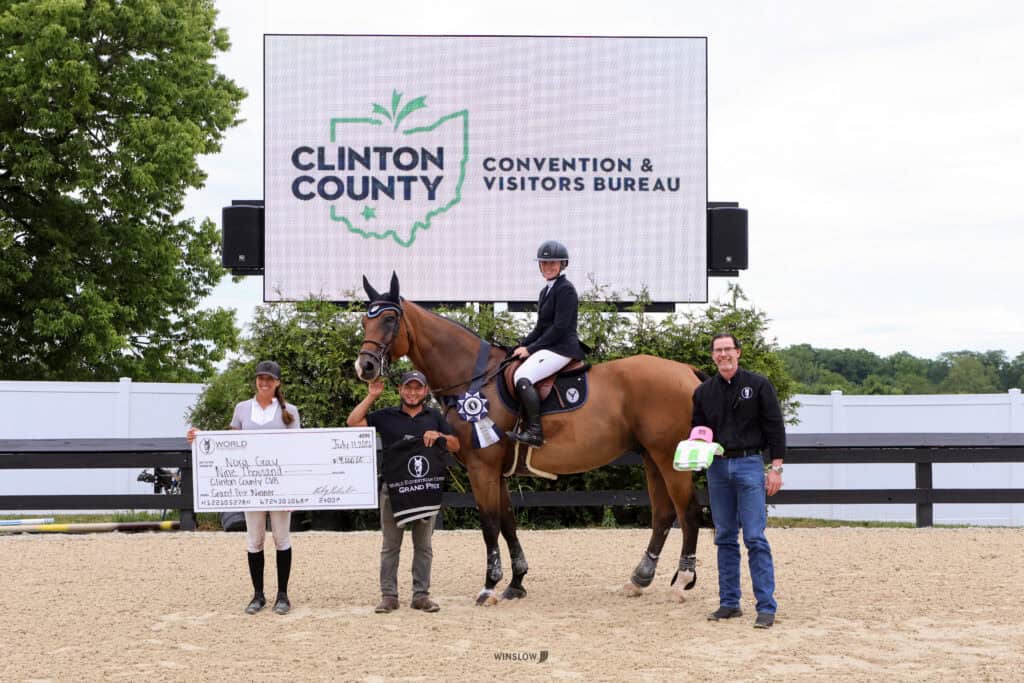 Four people and a horse pose in front of a Clinton County Convention & Visitors Bureau sign; one holds a large check, another person is mounted on the horse, and another holds a prize bucket.