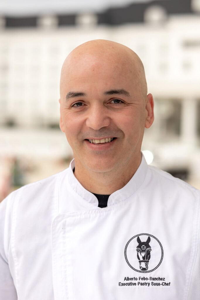 A man in a white chef’s coat with a logo and nametag stands outdoors, smiling at the camera. The background is blurred and shows a white building.