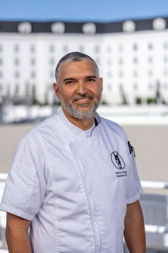 A chef in a white uniform stands outdoors, smiling at the camera, with a large white building and clear sky in the background.