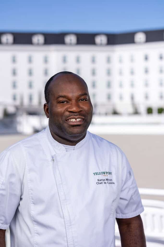 A man in a white chef’s coat stands outdoors, smiling, with a large white building in the background.