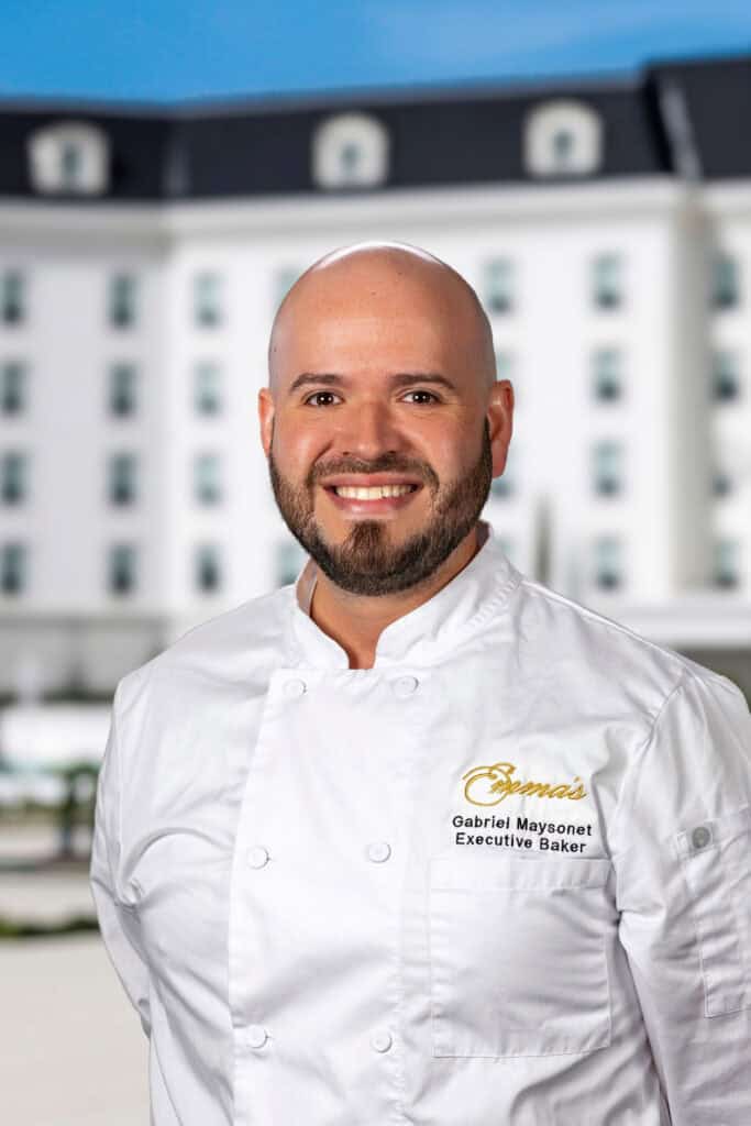 A man wearing a white chef’s coat stands outdoors, smiling, with a large white building in the background.