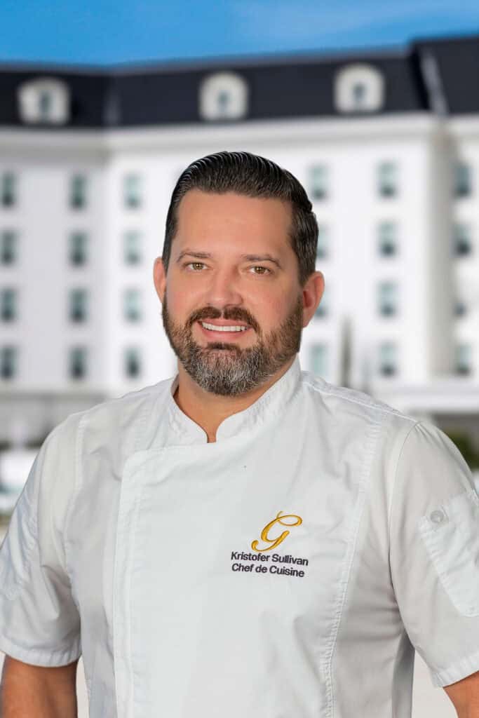 A chef in a white uniform stands outdoors in front of a large white building. The uniform reads 