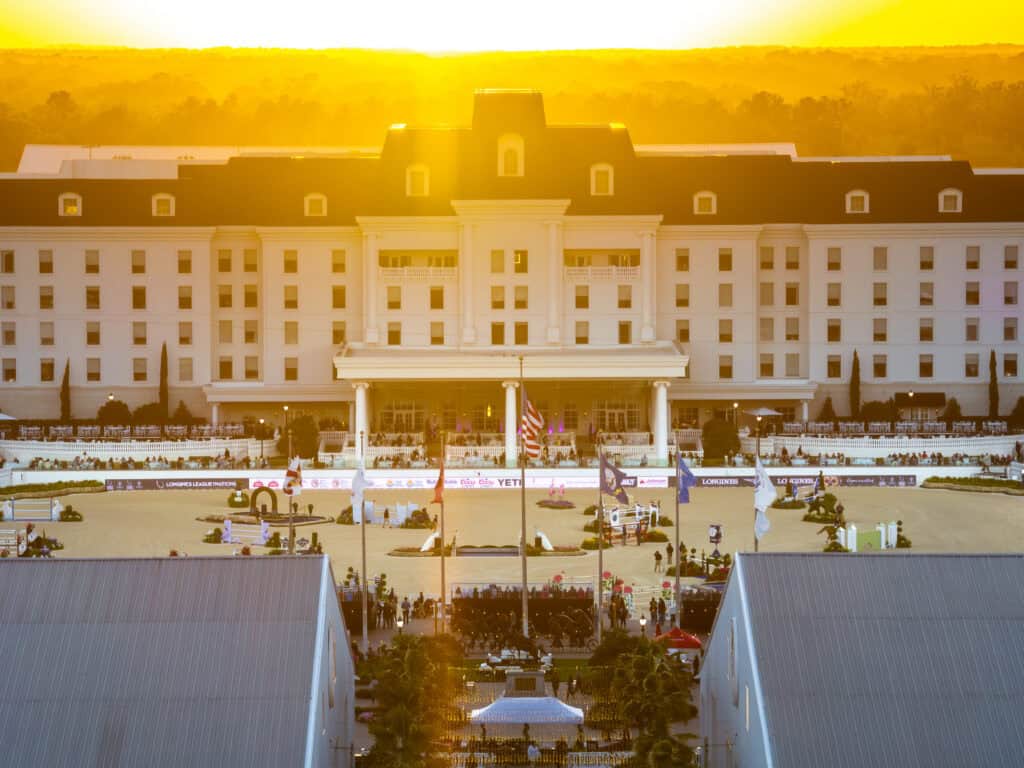 A large white hotel stands behind an equestrian arena at sunset, with flags, spectators, and event structures visible in the foreground.