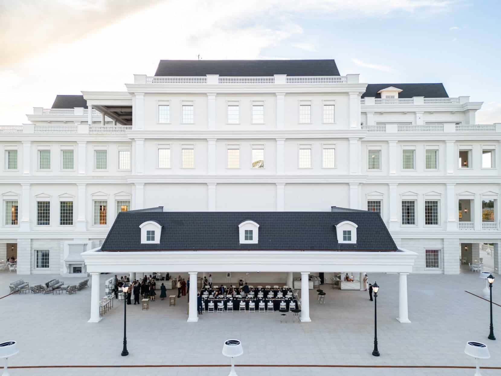 A group of people sits in rows under a pavilion in front of a large white classical-style building on a clear day.
