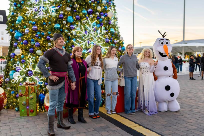 A group of people and two costumed characters stand in front of a decorated Christmas tree with lights, ornaments, and wrapped presents.
