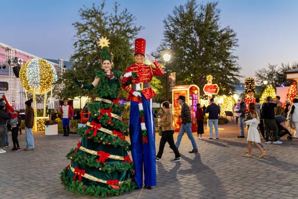 Two performers on stilts dressed as a Christmas tree and a toy soldier stand in a festive outdoor area with people and holiday lights in the background.