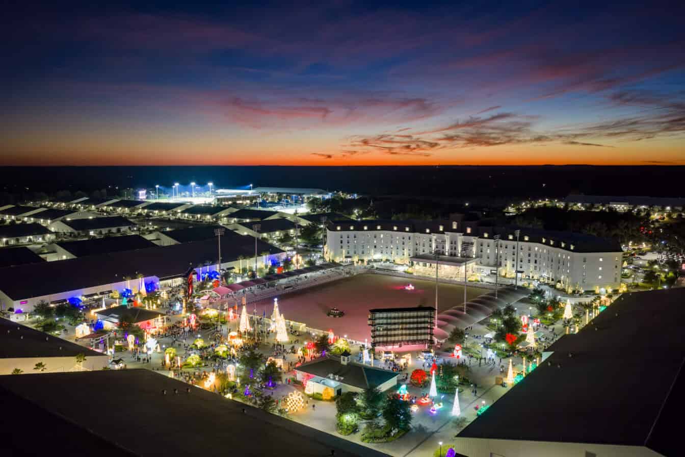 Aerial view of a brightly lit complex with holiday decorations at dusk, featuring a large central building and an outdoor plaza under a colorful sunset sky.