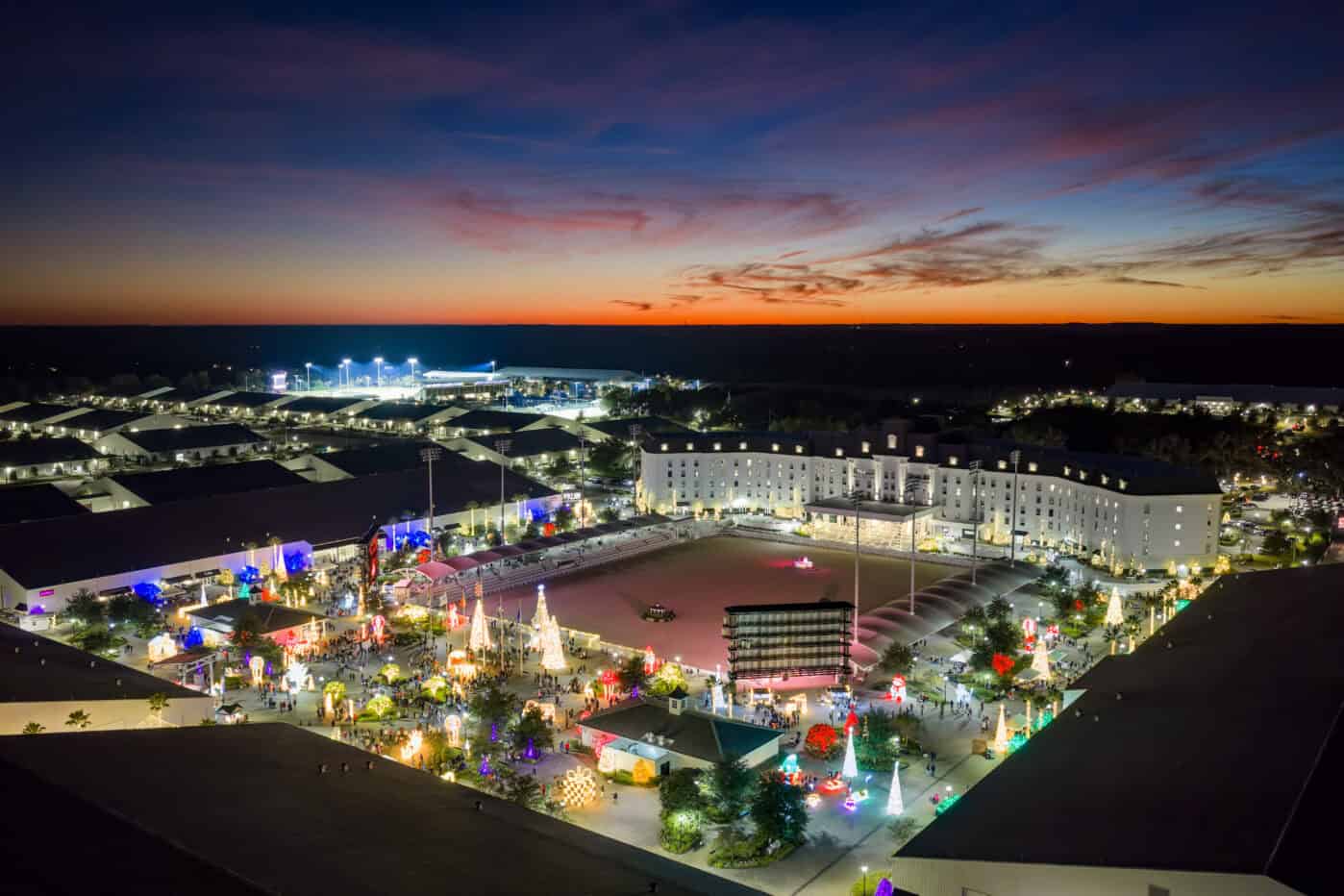 Aerial view of a brightly lit complex with holiday decorations at dusk, featuring a large central building and an outdoor plaza under a colorful sunset sky.