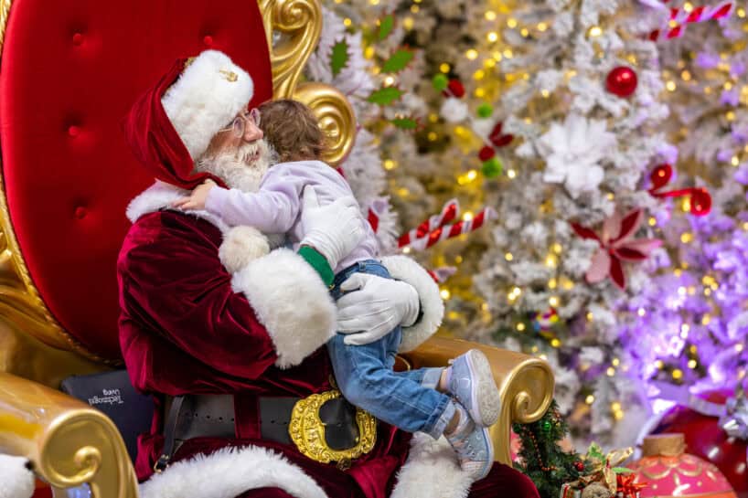 A child sits on Santa Claus’s lap, hugging him, with a decorated Christmas tree and festive ornaments in the background.