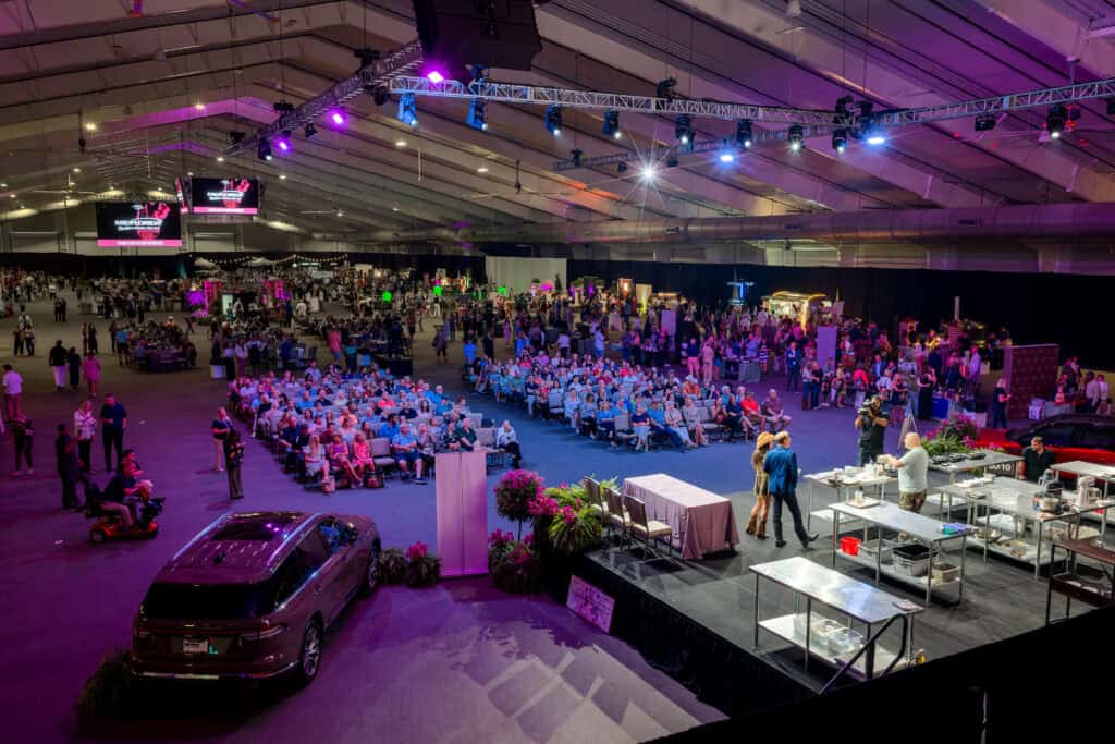 Large indoor event with audience seated facing a stage, where cooking demonstration takes place; car display and vendor booths visible under purple lighting.