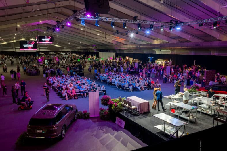Large indoor event with audience seated facing a stage, where cooking demonstration takes place; car display and vendor booths visible under purple lighting.