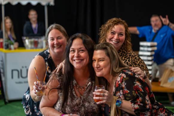 Four women smiling and posing for a photo at an indoor event, with drinks in hand; a man in the background makes a peace sign gesture.