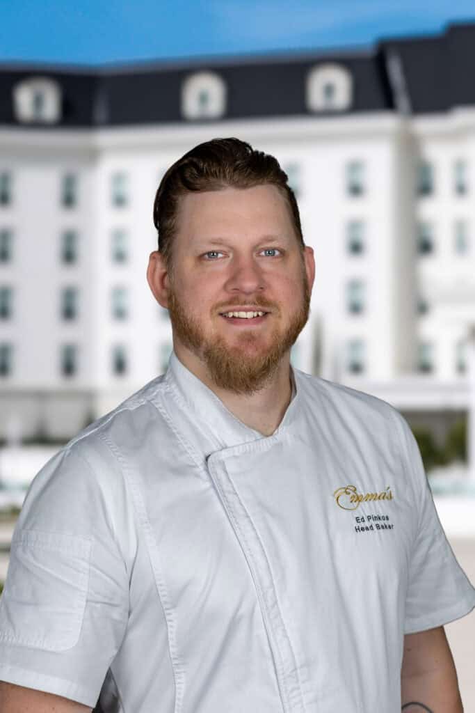 A man with a beard wearing a white chef’s coat stands in front of a large white building with many windows.