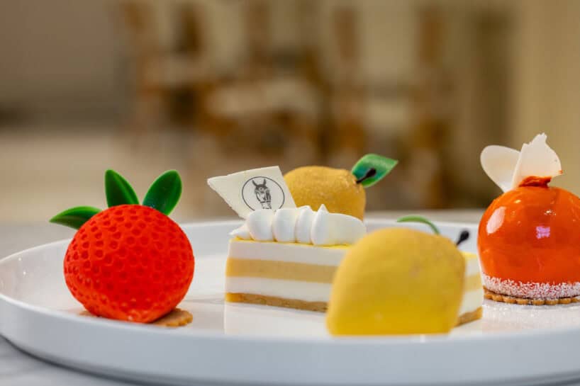 A white plate with four assorted elegant desserts, including a strawberry-shaped mousse, a lemon-shaped mousse, an orange-glazed sphere, and a layered cake with whipped topping.