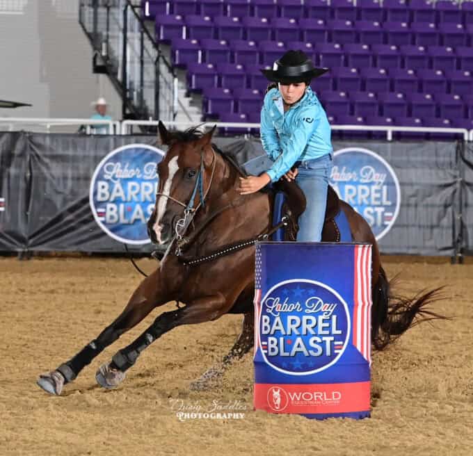 A rider in a blue shirt and black hat steers a horse sharply around a barrel at an indoor barrel racing event labeled "Labor Day Barrel Blast.
