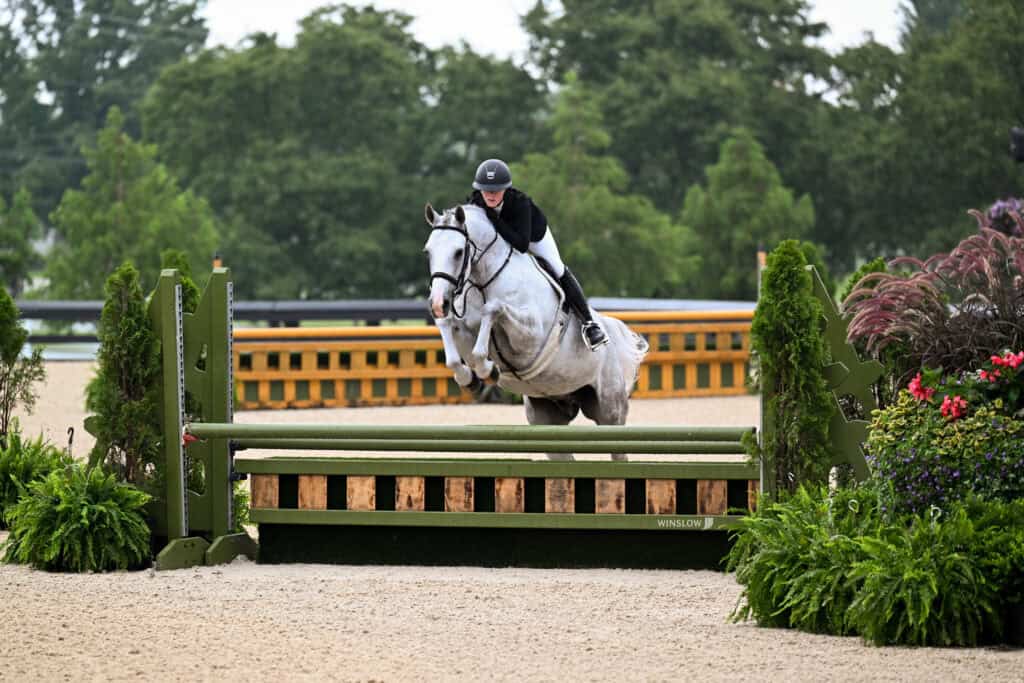 A rider in a helmet and jacket guides a gray horse over a green jump in an outdoor equestrian arena with trees in the background.