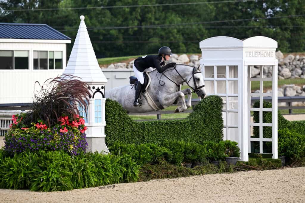 A rider on a gray horse jumps over an obstacle designed like a hedge and a telephone booth at an outdoor equestrian event.