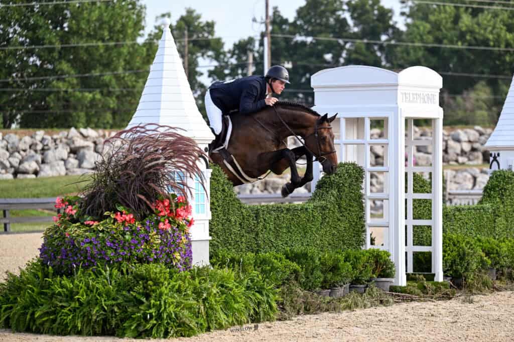 A horse and rider jump over a hedge obstacle decorated with plants and a white structure resembling a phone booth during an equestrian event.