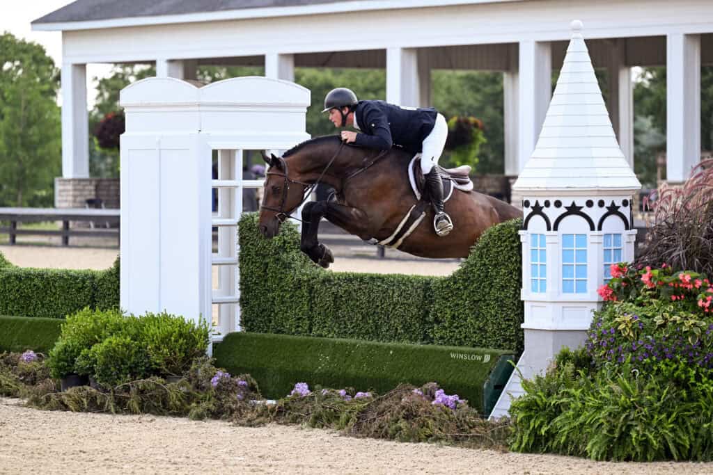 A rider in formal attire guides a horse over a green hedge jump during an equestrian event, with a white structure and plants in the background.