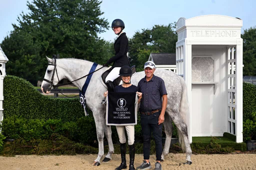 A rider on a white horse poses with two people holding a World Champion banner beside a white telephone booth outdoors.