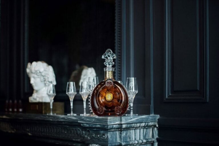 A crystal decanter filled with brown liquid is displayed on a marble surface, surrounded by four empty glasses. Marble busts are visible in the background against dark paneled walls.