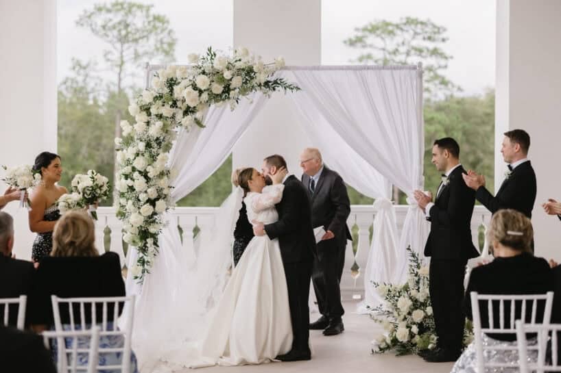 A bride and groom kiss under a white floral arch during their wedding ceremony as guests and attendants watch and applaud.