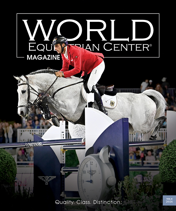 Equestrian rider in a red jacket jumps a white horse over an obstacle at a competition, featured on the cover of World Equestrian Center Magazine.