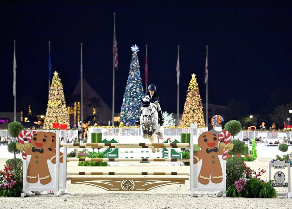 An equestrian jumps a horse over a festive obstacle decorated with gingerbread men, with illuminated Christmas trees and holiday decorations visible in the background at night.
