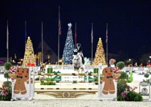 An equestrian jumps a horse over a festive obstacle decorated with gingerbread men, with illuminated Christmas trees and holiday decorations visible in the background at night.