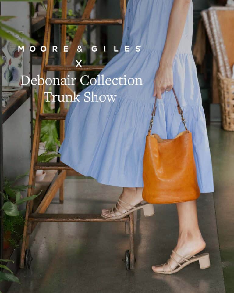 A person in a light blue dress and beige sandals stands near a wooden ladder, holding an orange leather bag. Promotional text for a trunk show appears over the image.