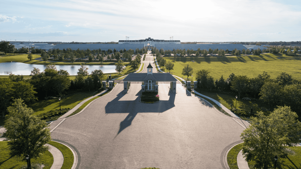 A wide, tree-lined driveway leads to a large, distant building with a central tower, flanked by ponds and lawns under a bright sky.