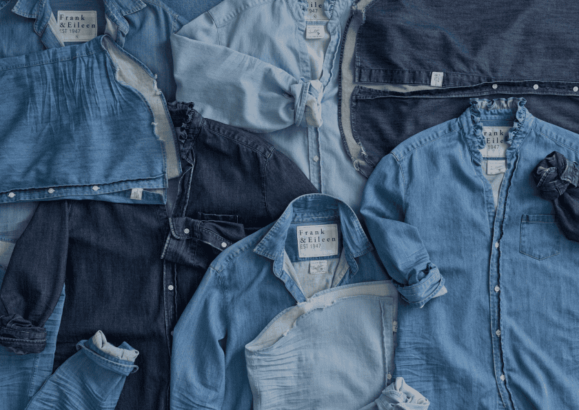 Several blue denim and chambray shirts, mostly labeled Frank & Eileen, are laid out flat overlapping each other in varying shades of blue.