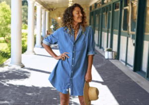 Woman standing outdoors in a blue denim shirt dress, holding a straw hat, and smiling, with columns and greenery in the background.