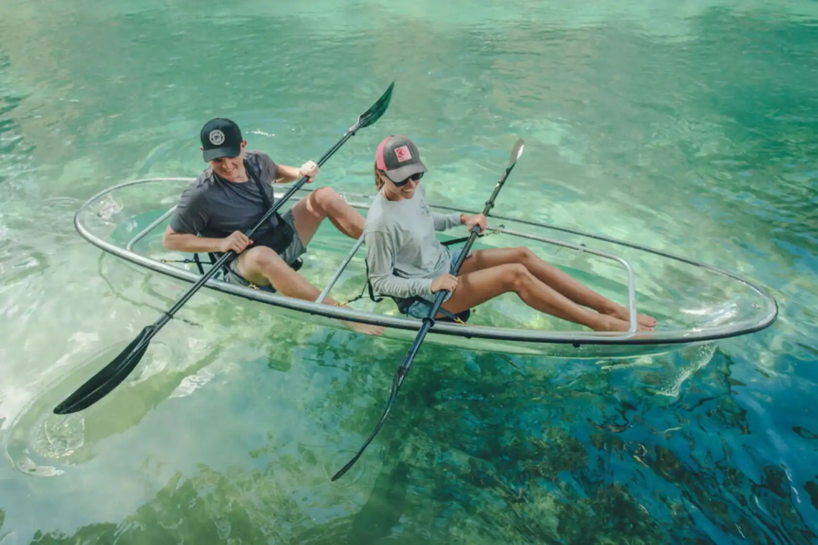 Two people paddle a transparent kayak over clear, turquoise water, revealing the underwater scenery below them.