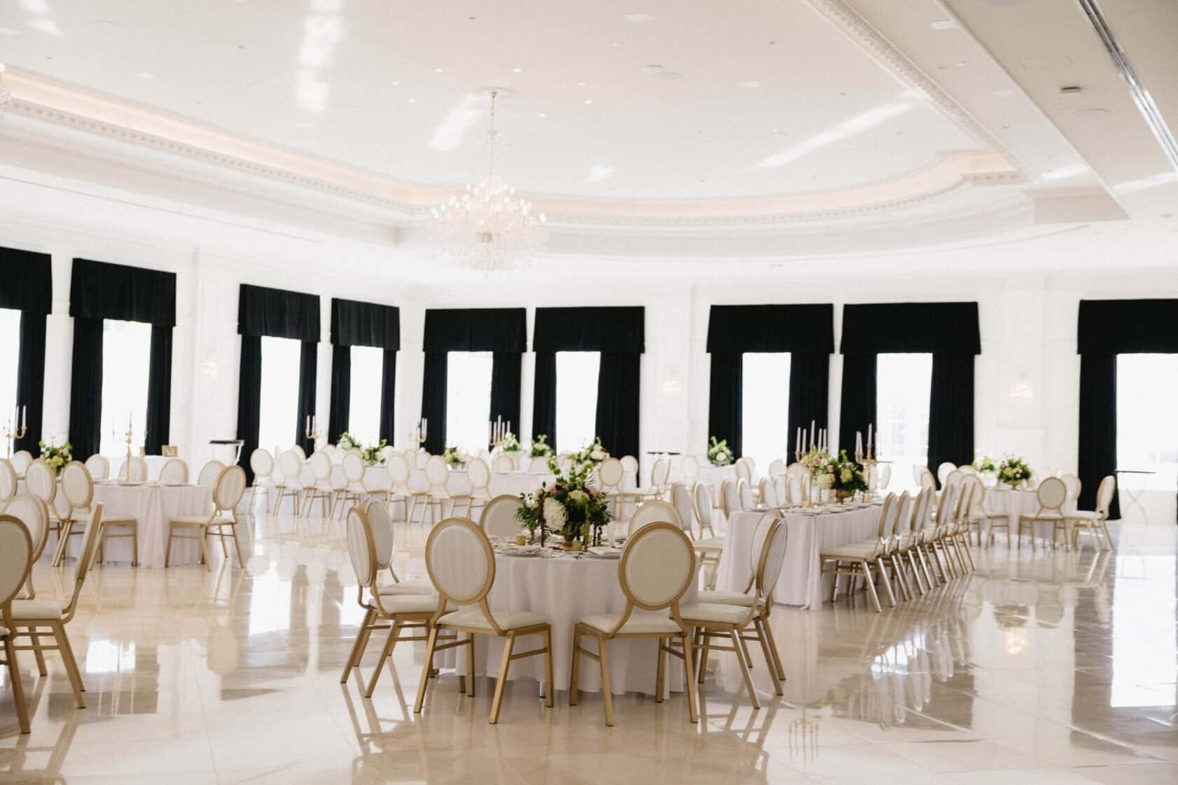 Elegant banquet hall with round tables, white chairs, black curtains, floral centerpieces, and a large chandelier. The room is bright with natural light and polished floors.