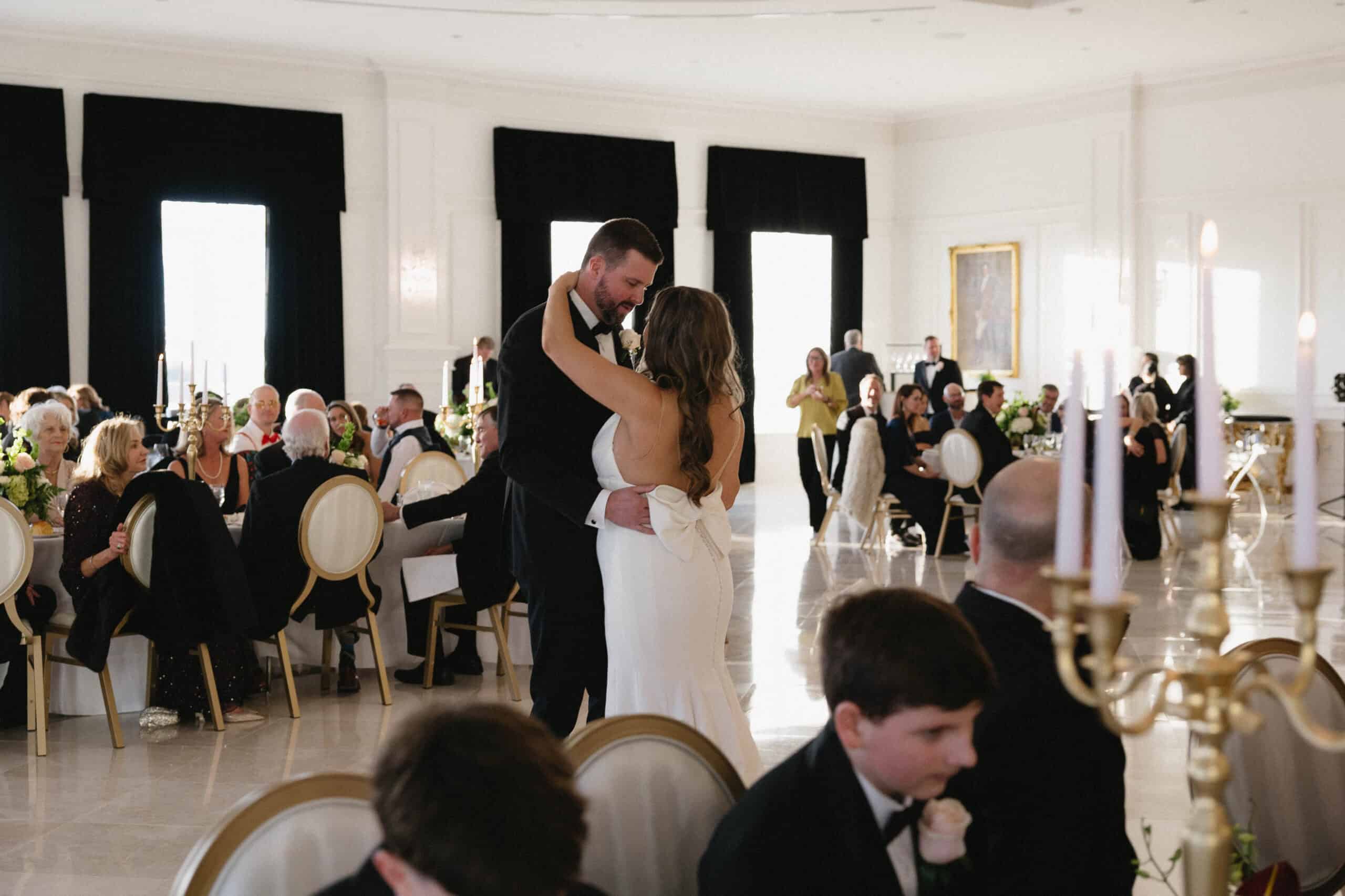 A bride and groom share their first dance in an elegant, well-lit reception hall as seated guests watch and dine at round tables.