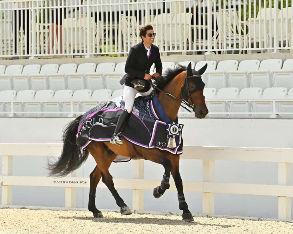 A rider in formal equestrian attire rides a horse wearing a decorative blanket and rosette in an arena with empty white seats.