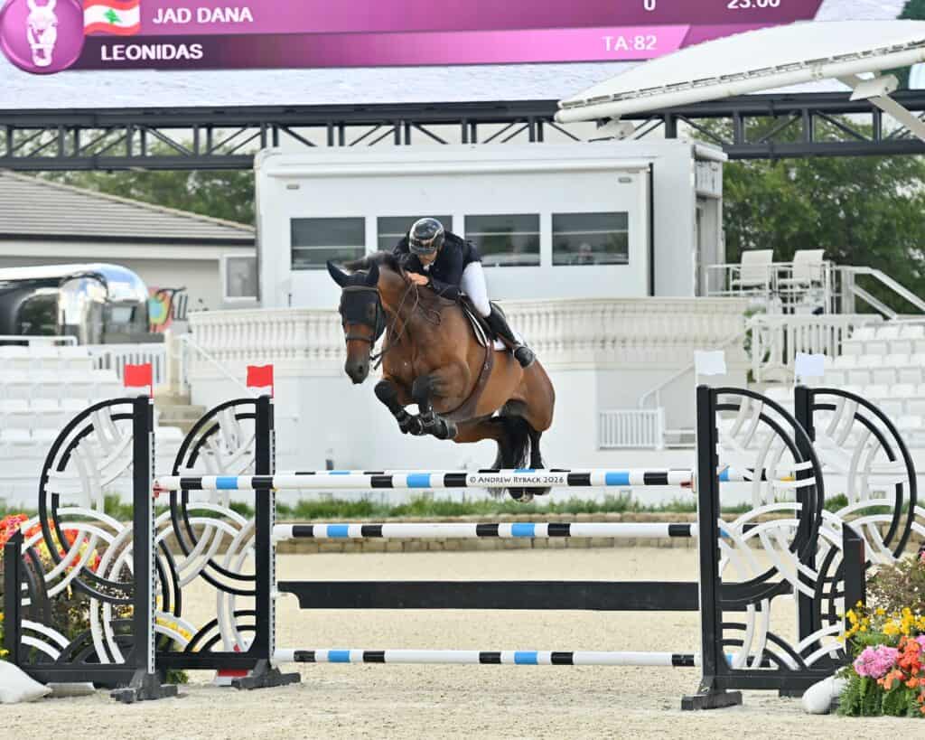 Equestrian rider and horse jump over an obstacle during a show jumping competition in an outdoor arena with empty seats visible in the background.